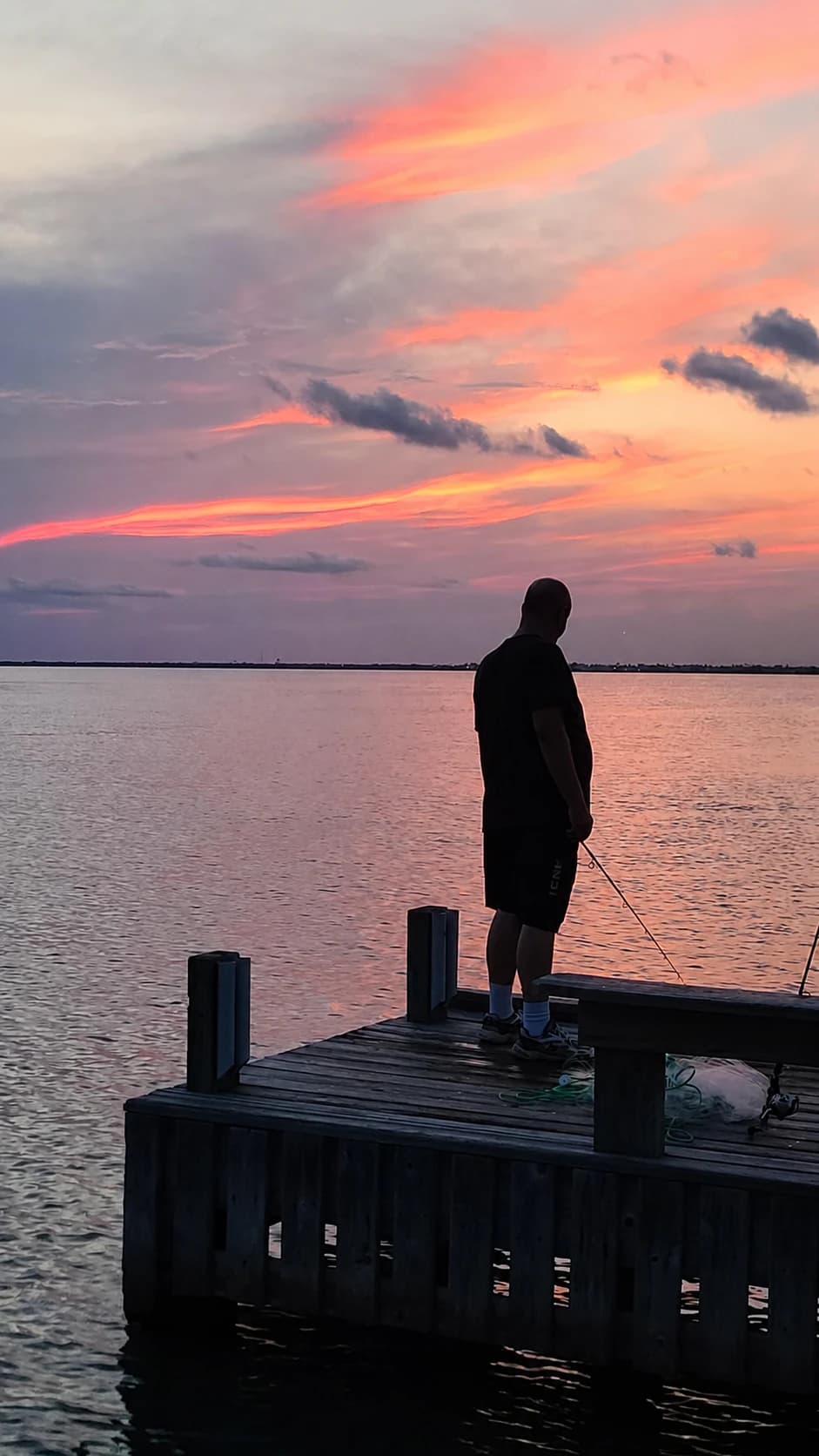 A distant silhouetted fisherman holds a rod from a wooden pier beneath a pink coastal sunset.