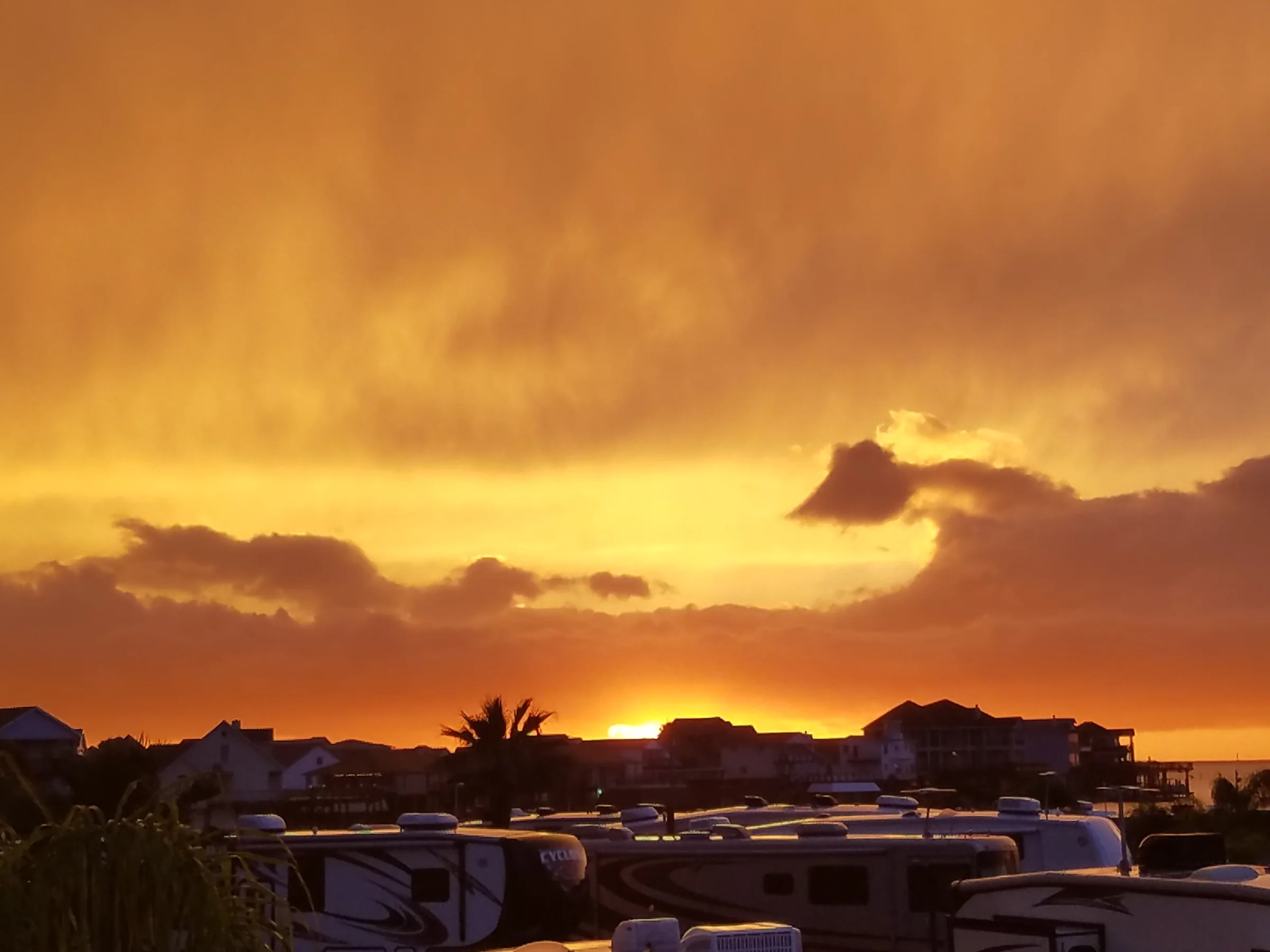 A warm orange sunset glows above the rooftops of RVs at the waterfront community.