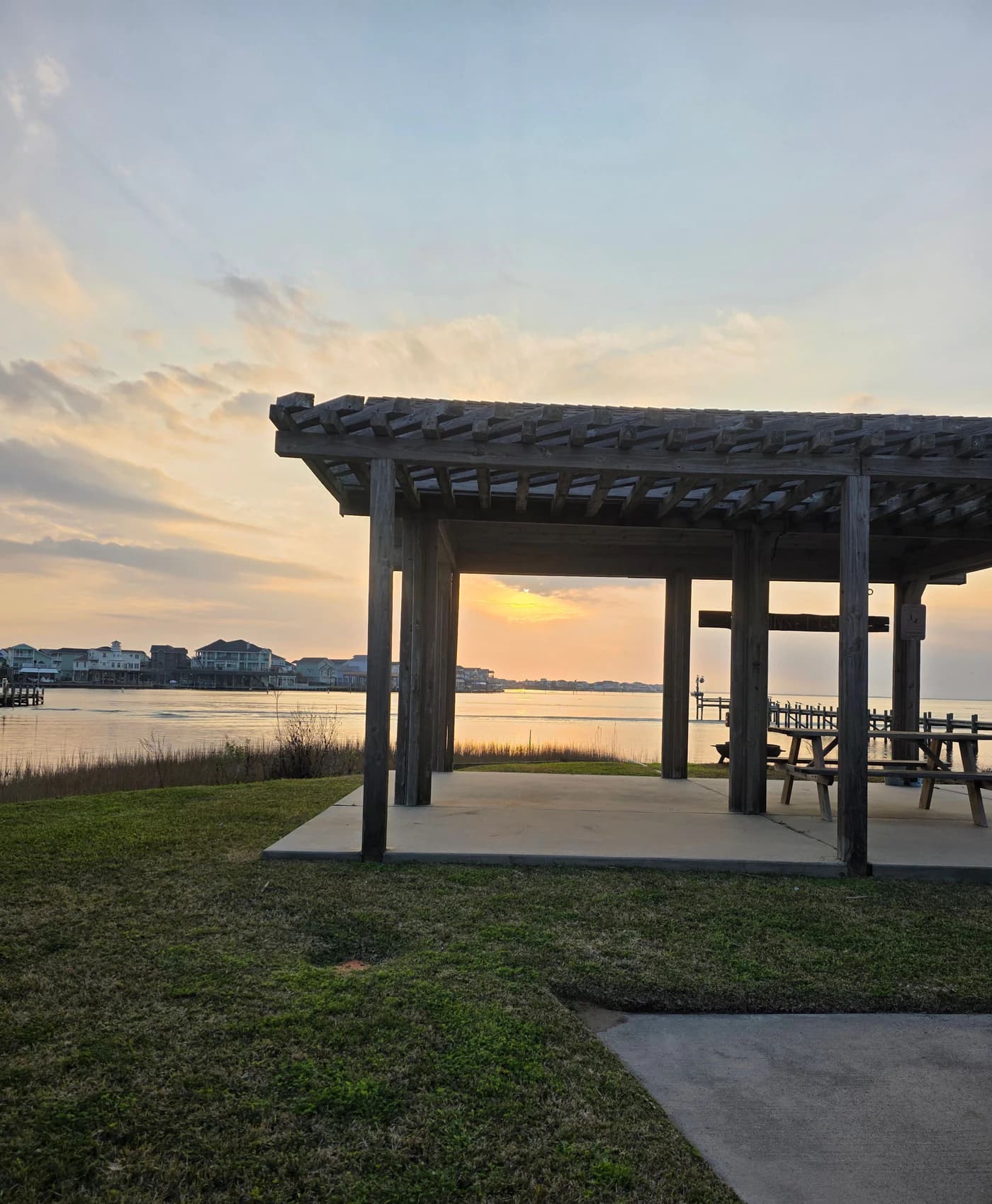A wooden shade structure with picnic tables stands beside a lawn overlooking calm bay water.